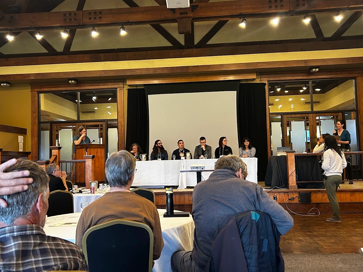 Youth and adult panelists seated at a long table on stage during a climate and education discussion at the NBEN Symbiosis conference, with audience members listening.