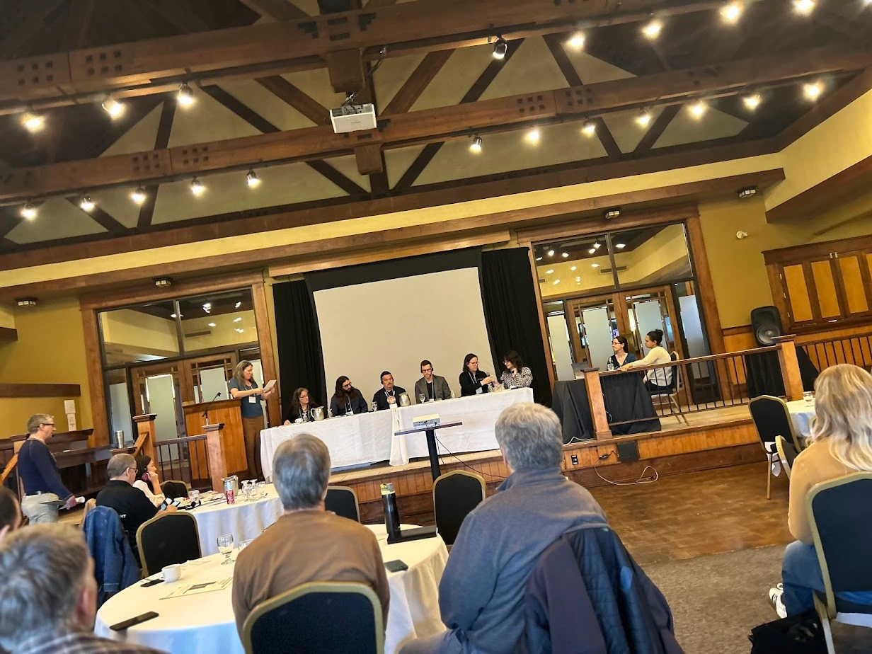 Youth and adult panelists seated on stage during a climate and education discussion at the NBEN Symbiosis conference, with attendees listening from round tables in a wood-beamed hall.