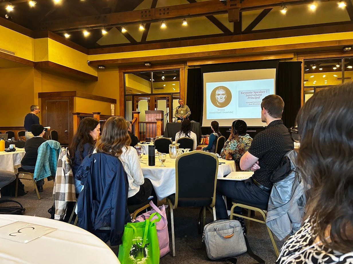 Speaker delivering a keynote address at the NBEN Symbiosis conference while attendees sit at round tables in a wood-beamed conference hall.