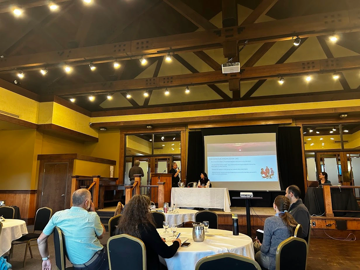 Conference presenter speaking beside a projected slide on Indigenous knowledge at the NBEN Symbiosis conference while attendees sit at round tables facing the stage.