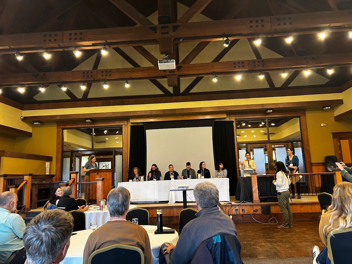 Panel discussion at the NBEN Symbiosis conference, with youth and adult panelists seated at a long table on stage, speaking to an audience of educators and environmental leaders in a wood-beamed conference hall.