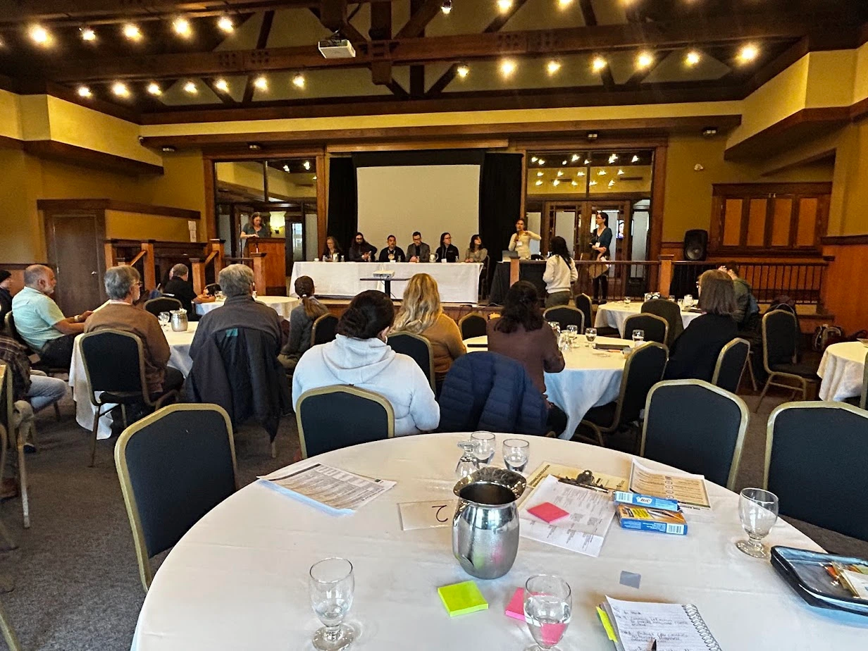 Wide view of the NBEN Symbiosis conference showing attendees seated at round tables facing a stage where a mixed youth and adult panel discusses climate action and environmental education.